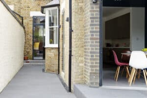 A modern kitchen with colorful chairs is visible through the bifold doors of a brick house, leading to a narrow courtyard, showcasing a stylish extension.