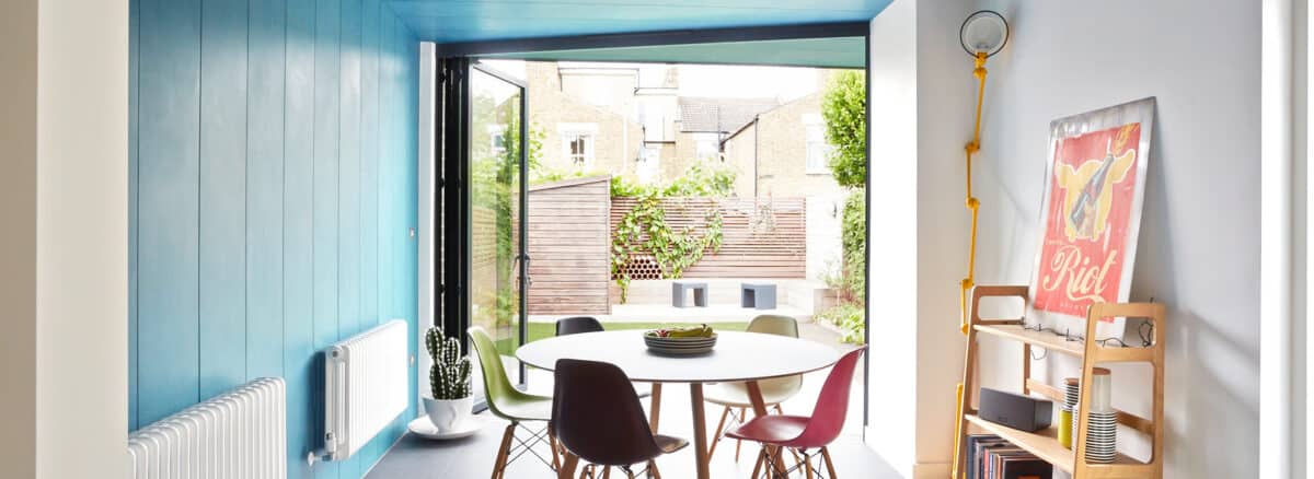 Bright dining area with a round white table, four chairs, blue accent wall and ceiling, bifold doors extension opening to a garden, and a shelf with books and a red poster.
