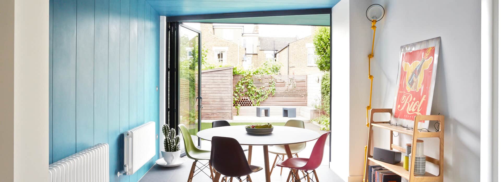 Bright dining area with a round white table, four chairs, blue accent wall and ceiling, bifold doors extension opening to a garden, and a shelf with books and a red poster.
