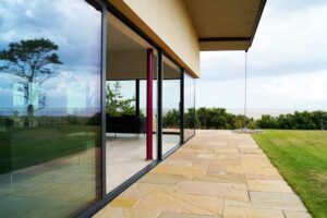 Modern house with expansive picture windows overlooking a grassy yard and trees, offering a stunning view of the distant sea under a cloudy sky.