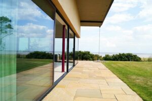 Modern house with expansive picture windows overlooking a grassy yard and distant sea view on a sunny day.