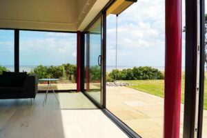 A modern interior with large picture windows and glass doors opens to a patio and grassy area, offering stunning views of the sea on a clear day.