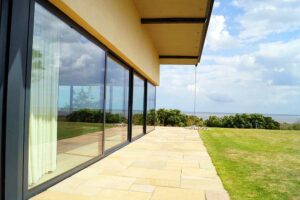 Modern house with expansive picture windows, overlooking a green lawn and ocean view.
