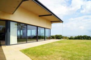 A modern house with expansive picture windows overlooks a grassy lawn and a distant ocean view under a partly cloudy sky.
