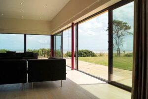 The interior of a modern room features large picture windows and glass sliding doors opening to a patio, grassy area, and a view of the sea under a partly cloudy sky. Two black sofas are visible in the room.