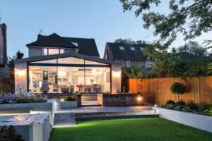 Modern house with large glass windows and sliding doors at dusk, overlooking a landscaped garden with a lawn, stepping stones, and a wooden fence.