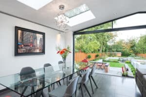 A modern dining room features a glass table and gray chairs beneath a chandelier, with large sliding doors revealing a lush garden view. A painting tastefully adorns the wall.