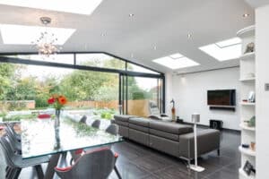 Modern living room with a glass dining table, gray sofas, and a wall-mounted TV. Large windows and skylights flood the space with light. Large sliding doors open to reveal a stunning garden view, seamlessly blending indoor and outdoor living.