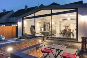 Modern home with large glass windows and large sliding doors, showing a well-lit interior with a dining area and living space. Outdoor patio features a small table and chairs, with surrounding brickwork and lighting.