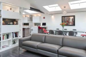 Modern living room with a gray sofa, wall shelves, and an open kitchen. A glass dining table with red chairs is under skylights. Large sliding doors let in ample light, complementing the cityscape painting on the white wall.