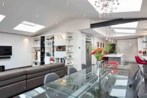 Modern open-plan living space featuring large sliding doors, a glass dining table, leather sofa, wall-mounted TV, bookshelves, and a white kitchen with an island and bar stools under skylights.