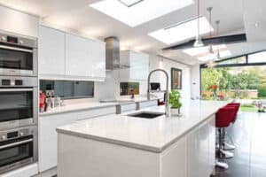 Modern kitchen with a white island, integrated appliances, red bar stools, and skylights. Large sliding doors and large windows open to a garden view.