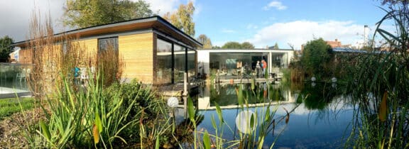 Modern building with large glass windows and ultra slim sliding doors, overlooking a pond surrounded by reeds and plants.