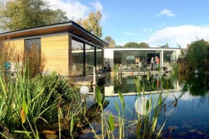 A modern house with ultra slim sliding doors features large glass windows beside a pond, surrounded by tall grasses and vegetation under a partly cloudy sky. Two people stand on a wooden deck over the water.