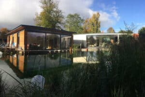 Modern house with ultra slim sliding doors and large glass windows next to a tranquil pond, surrounded by trees and grass under a partly cloudy sky.