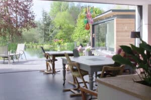 Dining area with wooden tables and chairs, offering a view of the garden and pond through ultra slim sliding doors. Colorful pendant lights hang from the ceiling.