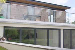 Modern two-story house with large glass windows and ultra slim sliding doors leading to a balcony featuring outdoor furniture.
