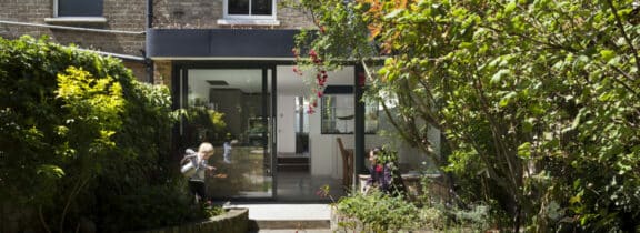 A garden with lush greenery and a person watering plants. A glass sliding door beneath a sleek flat roof extension with overhang leads to the interior of a house.