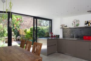 Modern kitchen with large glass doors opening to a garden, complemented by a flat roof extension with overhang. Wooden dining table with chairs, sleek countertops, and kitchen appliances create an inviting space. Bright greenery visible outside enhances the warm ambiance.