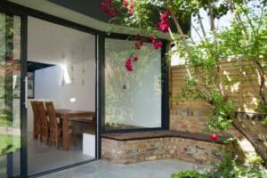 Seen through large glass doors, the modern dining area features a wooden table and chairs. A flat roof extension with overhang shelters this space, while the brick exterior showcases blooming plants and a wooden fence in the background.