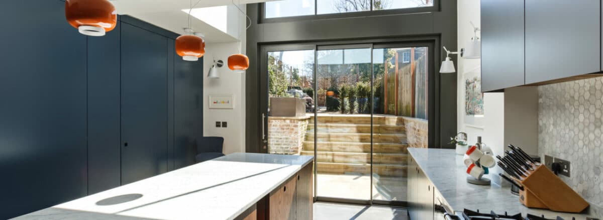 Modern kitchen with fixed windows, marble countertops, blue cabinets, and orange pendant lights. Glass doors open to a sunny patio with wooden stairs leading to a garden.