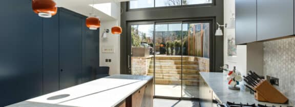 Modern kitchen with fixed windows, marble countertops, blue cabinets, and orange pendant lights. Glass doors open to a sunny patio with wooden stairs leading to a garden.