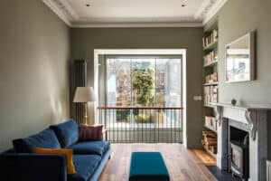 A living room with a blue sofa, a marble fireplace, and bookshelves sits beneath a flat roof extension with overhang. Sunlight pours through the large window overlooking a garden.