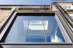 Low-angle view of a modern building featuring a large, reflective glass window and partial brick facade, complemented by a stylish flat roof extension with overhang against the clear blue sky.