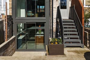 Modern flat roof extension with an overhang, featuring large glass windows and a brick exterior. A staircase leads up to a door, complemented by a nearby planter brimming with flowers.