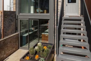 A modern brick house exterior featuring large glass windows, a sleek metal staircase, and a small flower garden is complemented by a flat roof extension with an overhang.