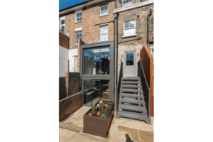 A modern two-story home extension featuring large fixed windows, outdoor stairs, and a small garden bed in front.