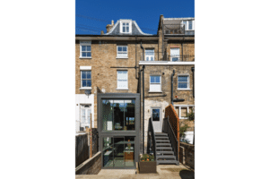 A brick townhouse with a glass extension at the back, featuring a flat roof extension with an overhang and large window showcasing visible interiors. A staircase leads to the upper levels.