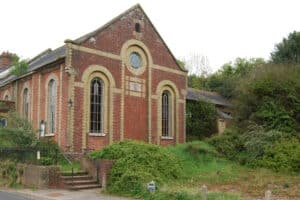 A brick building with arched windows and a circular window above the door, surrounded by overgrown grass and bushes next to a road.