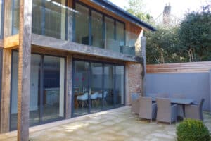 Modern two-story house with large glass windows, wooden and brick exterior. Outdoor patio with wicker dining set on a stone-paved surface.