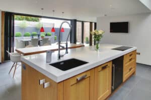 Modern kitchen with a large island featuring a sink and stovetop. Sliding doors open to a patio with a dining set and red pendant lights.