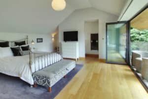 Spacious bedroom with a large bed, a television on a dresser, and an open folding door leading to a balcony with seating. Light wooden flooring and a patterned bench complete the decor.