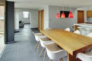 Modern dining area with a wooden table and white chairs, adjacent to a kitchen with red pendant lights. A glass wall opens to a patio, and a hallway leads to another room with a couch.