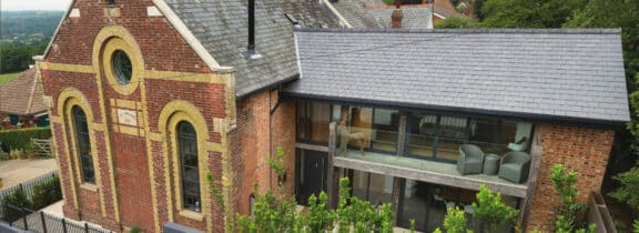 A modern home with a large glass section merges seamlessly with a historic brick building, featuring a round window and arched details.
