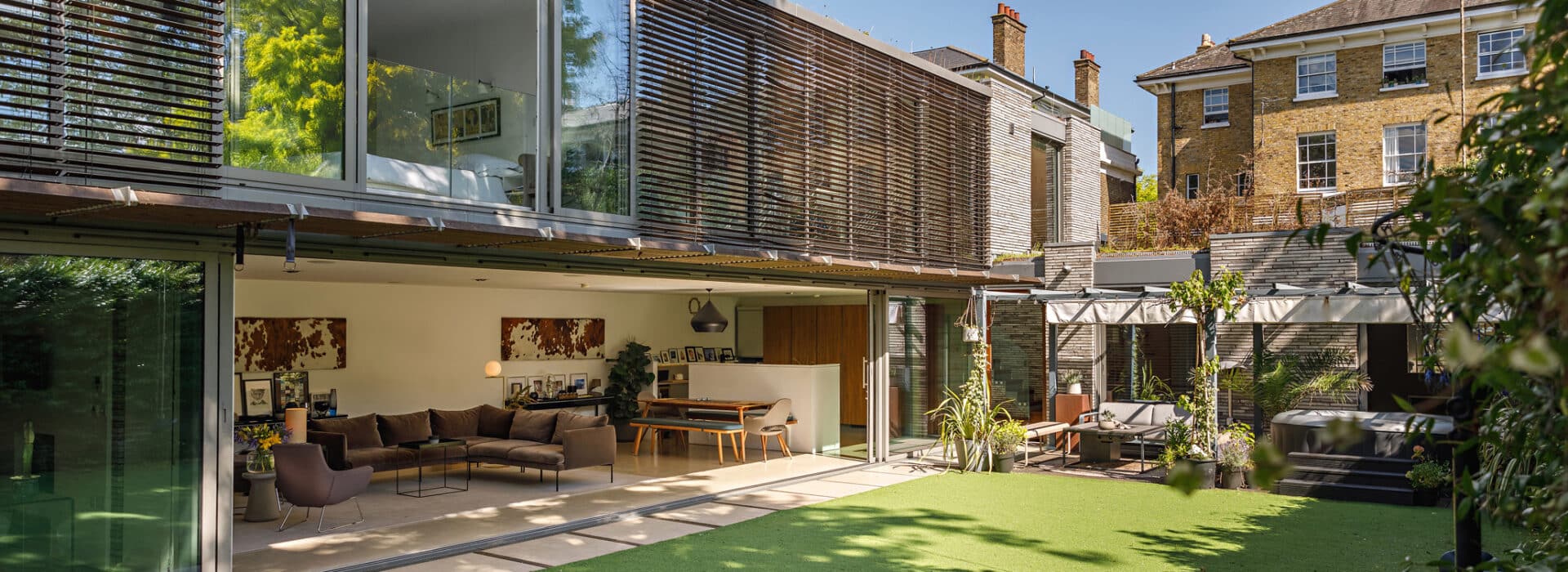 Modern house with large glass sliding doors open to a patio and artificial grass lawn, revealing a living area with contemporary furnishings.