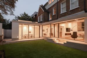 Modern house with large glass windows and sliding aluminium doors, offering a well-lit interior, viewed from a lawn in the garden area.