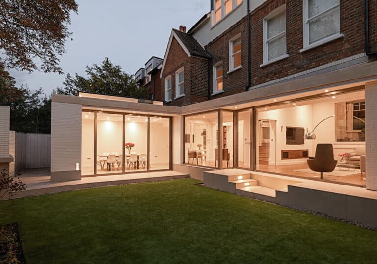 Modern house with large glass windows and sliding aluminium doors, offering a well-lit interior, viewed from a lawn in the garden area.