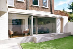 Modern extension with large glass windows and sliding aluminium doors, featuring a brick exterior and a small step leading to a grassy backyard. Indoor seating is visible through the windows, offering a seamless blend of indoor and outdoor living spaces.
