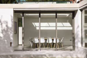 Exterior view of a modern dining room with large, sliding aluminium doors, showing a table surrounded by white chairs and a ceiling shadow pattern.