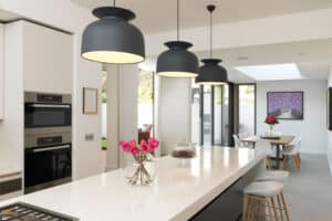 Modern kitchen with a large island, pendant lights, built-in ovens, and glass vase with pink flowers. Sliding aluminium doors lead to a dining area featuring a table and chairs in the background.