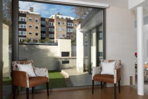 Two brown chairs with white cushions face a large window paired with sliding aluminium doors, overlooking a patio and apartment buildings.
