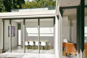 Modern dining area with a table and white chairs, viewed through large sliding aluminium doors. An adjacent room features a brown armchair, while trees are visible outside.