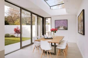 Modern dining room with a wooden table, white chairs, a vase of pink flowers, and sliding aluminium doors opening to a garden. A large skylight and floral artwork enhance the space.