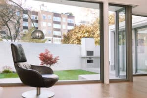 Modern living room with a black swivel chair, sliding aluminium doors opening to large glass windows, and a view of a small garden featuring a red-leafed tree and white brick barbecue area.
