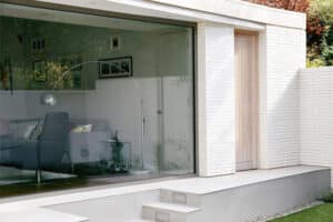 Modern house with large sliding aluminium doors, a white brick exterior, and a three-step entrance leading to a green lawn. The interior is visible with a sofa and floor lamp.