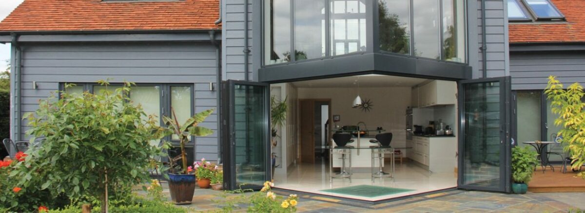 Modern house with a red-tiled roof features large glass doors opening to a patio, offering a glimpse of the kitchen inside. An extension with a glass roof allows natural light to flood the space, while the plant-filled garden enhances its charm.